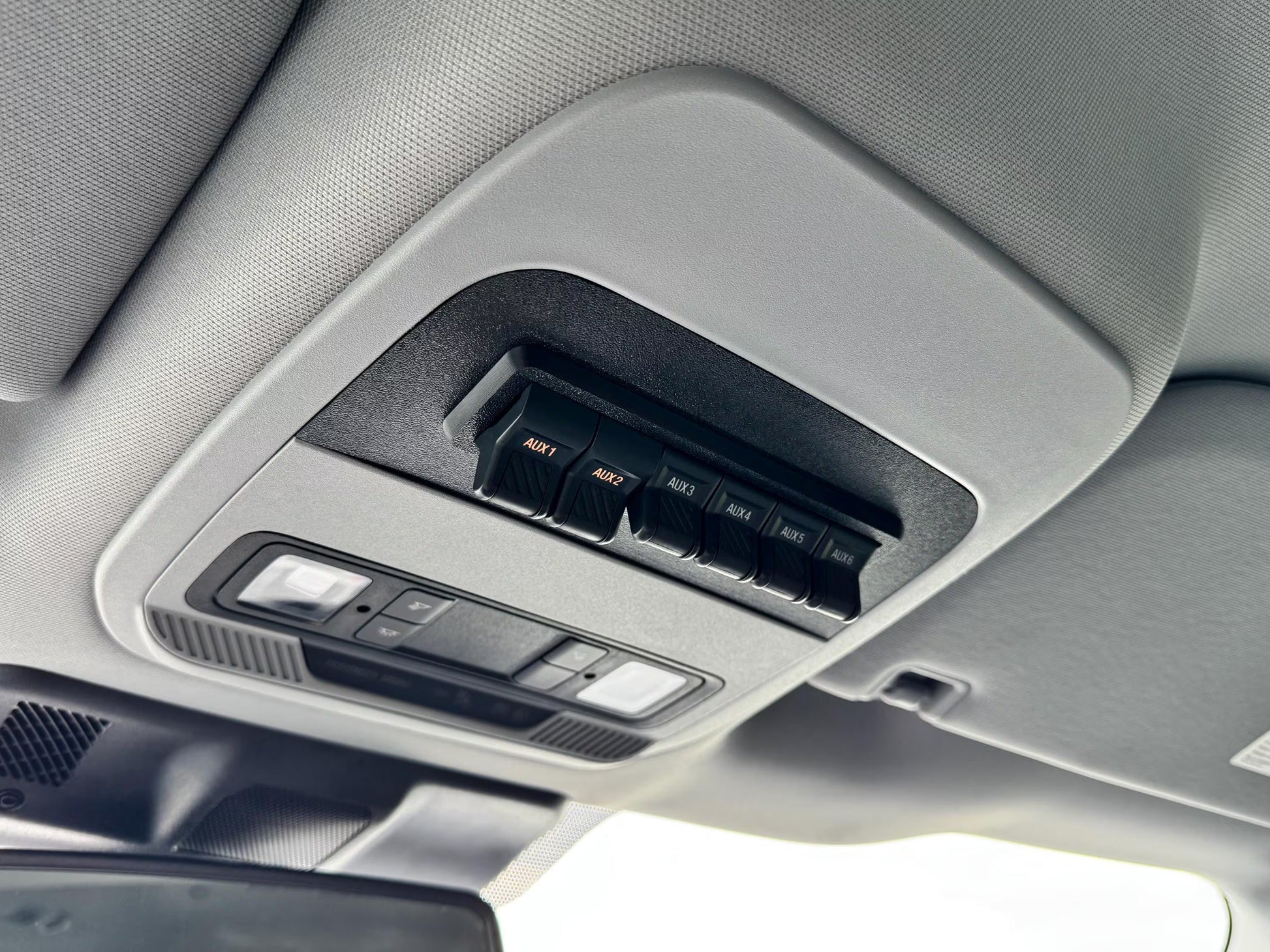 Close-up of a car interior overhead console with sun visor controls, including black aux switch buttons labeled and a light control module, mounted on a gray fabric headliner.