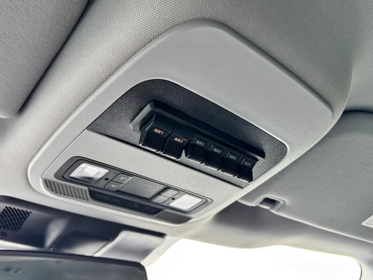 Close-up of a car interior overhead console with sun visor controls, including black aux switch buttons labeled and a light control module, mounted on a gray fabric headliner.