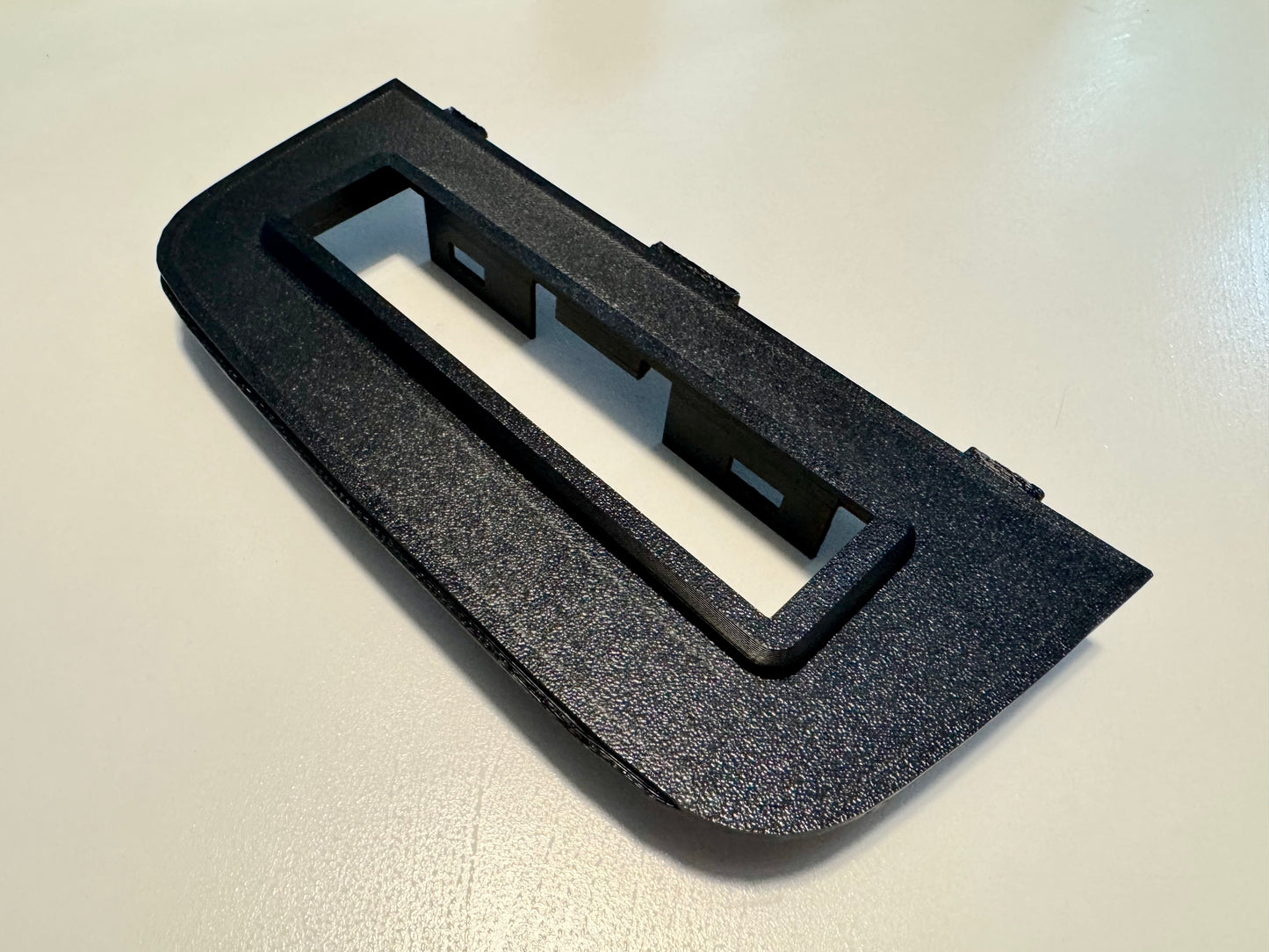 A black textured plastic dashboard trim piece with a rectangular cutout, photographed on a light off-white surface.