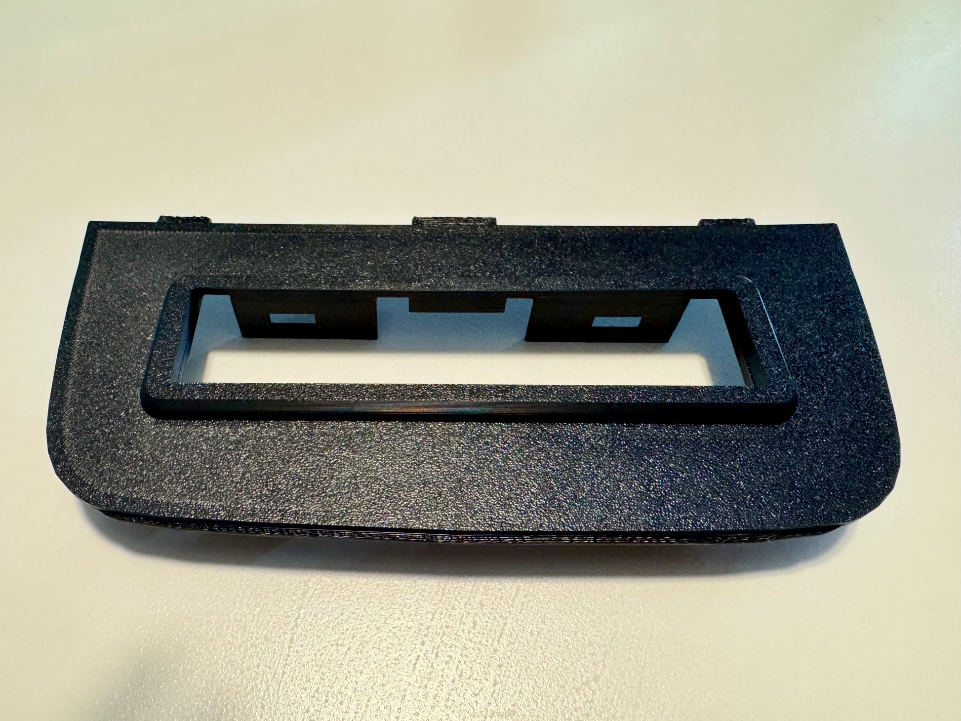 A black textured plastic dashboard trim piece with a rectangular cutout, photographed on a light off-white surface.